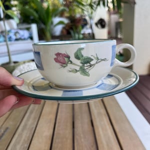 May include: A cream-coloured teacup and saucer featuring a rose motif. The edges of the cup and saucer are adorned with a blue and white gingham pattern. The teacup is held by a person.