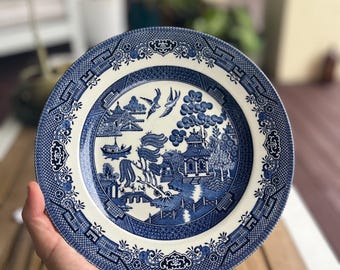 May include: Blue and white ceramic plate with a Willow pattern design. The plate features a central landscape scene with a bridge, buildings, and birds, surrounded by a decorative border. The plate is held over a wooden surface.