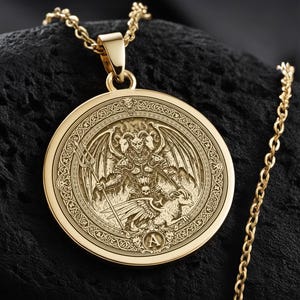 May include: Gold-colored pendant necklace featuring a detailed engraved design of a winged creature with a trident. The pendant is circular and hangs from a delicate chain. The necklace rests on a dark, textured surface, creating a striking contrast.
