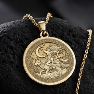 May include: A gold-colored pendant necklace featuring a detailed relief of a figure riding a wolf, with a crescent moon and stars. The round pendant hangs from a delicate chain. The necklace is displayed on a black stone.