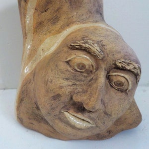 May include: A ceramic sculpture of a human face with a tilted head. The sculpture is primarily brown with lighter tan and white accents. The face has detailed features including eyes, eyebrows, and lips. The piece has a rough, textured surface.