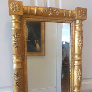 May include: A rectangular gold-colored mirror with ornate detailing. The frame features decorative carvings, including grape clusters and floral patterns. The mirror reflects a partial view of a room and a framed painting.