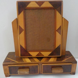 May include: A wooden vanity mirror with a rectangular frame featuring light and dark brown geometric wood inlay patterns. The mirror is mounted on a rectangular base that includes two small drawers, each equipped with a simple wooden handle.