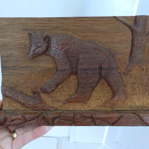 May include: A brown wooden relief carving of a bear walking to the left. The bear is carved in detail, with fur and facial features visible. A tree and a log are also carved into the wooden panel. The base of the carving has a stone-like texture.