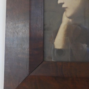 K&ouml;nnte beinhalten: Ein dunkelbrauner Holzbildrahmen mit einem Schwarzwei&szlig;foto einer Frau darin. Die Frau tr&auml;gt einen dunklen Hut und hat ihr Kinn auf ihrer Hand ruhen.