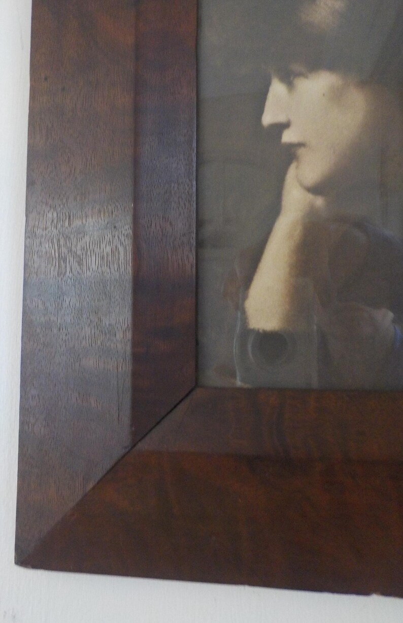 K&ouml;nnte beinhalten: Ein dunkelbrauner Holzbildrahmen mit einem Schwarzwei&szlig;foto einer Frau darin. Die Frau tr&auml;gt einen dunklen Hut und hat ihr Kinn auf ihrer Hand ruhen.