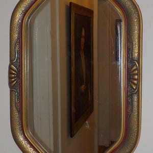 May include: Oval-shaped mirror with a decorative gold frame. The frame has a textured surface with red and blue accents. The mirror reflects a painting and a white wall.