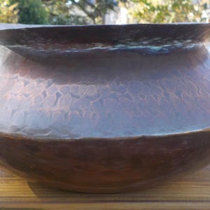 May include: A large, hammered copper planter with a wide, rounded base and a slightly flared rim. The planter has a rustic, aged appearance with a mottled brown and copper patina.