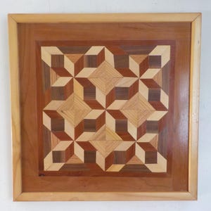 May include: A square wooden artwork with a geometric pattern. The design features a repeating pattern of three-dimensional cubes and star-like shapes, crafted from various shades of wood, including light and dark browns, and beige. The artwork is framed in a light brown wood.