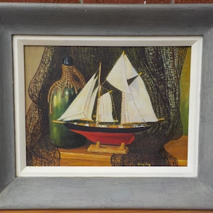 May include: A framed painting depicts a sailboat with white sails, a red hull, and a black deck. A green bottle with a rope-like design and a fishing net are in the background. The painting is in a gray frame.