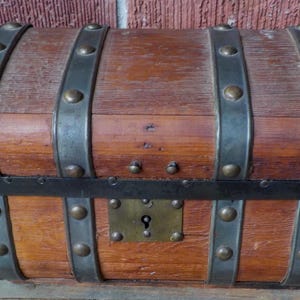 May include: A wooden chest with metal bands and rivets. The chest has a rectangular shape with a curved top and is secured with a metal lock. The wood has a warm, reddish-brown tone, and the metal bands are dark gray.