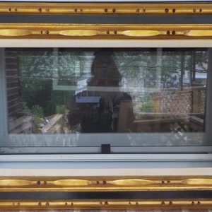May include: Ornate rectangular picture frame with a gold and black border. The frame surrounds a window, reflecting a backyard scene with trees and a deck. The frame has a decorative pattern and is mounted on a brick wall.