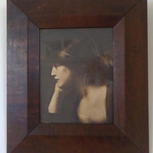 K&ouml;nnte beinhalten: Ein gerahmtes Sepia-Foto einer Frau, die den Kopf gesenkt hat und zur Seite schaut. Die Frau tr&auml;gt ein dunkles Kleid und hat ihre Haare nach hinten gebunden.