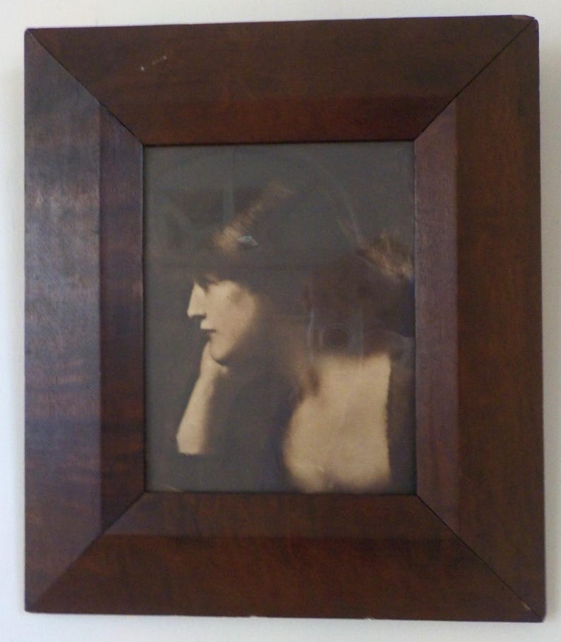 K&ouml;nnte beinhalten: Ein gerahmtes Sepia-Foto einer Frau, die den Kopf gesenkt hat und zur Seite schaut. Die Frau tr&auml;gt ein dunkles Kleid und hat ihre Haare nach hinten gebunden.