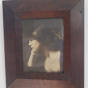 K&ouml;nnte beinhalten: Ein gerahmtes Sepia-Foto einer Frau, die ihren Kopf auf ihre Hand gelegt hat. Die Frau schaut zur Seite und hat dunkles Haar. Der Rahmen ist aus dunklem Holz.