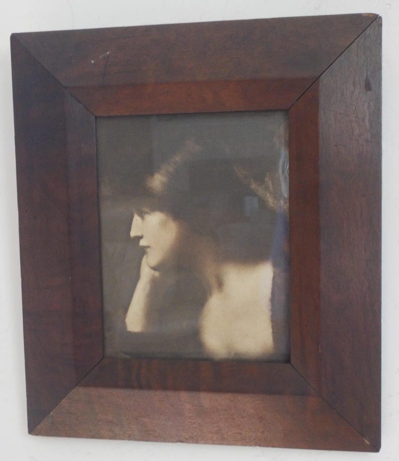 K&ouml;nnte beinhalten: Ein gerahmtes Sepia-Foto einer Frau, die ihren Kopf auf ihre Hand gelegt hat. Die Frau schaut zur Seite und hat dunkles Haar. Der Rahmen ist aus dunklem Holz.