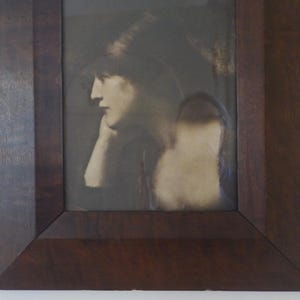 K&ouml;nnte beinhalten: Ein Schwarzwei&szlig;portr&auml;t einer Frau in einem dunklen Hut, die nach unten und nach links schaut. Das Bild ist in einem dunklen Holzrahmen gerahmt.