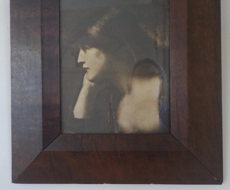 K&ouml;nnte beinhalten: Ein Schwarzwei&szlig;portr&auml;t einer Frau in einem dunklen Hut, die nach unten und nach links schaut. Das Bild ist in einem dunklen Holzrahmen gerahmt.