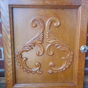 May include: Wooden cabinet door featuring an elaborate, carved floral pattern. The door is a warm, medium brown with a clear glass knob. The carved details are highlighted by the wood's natural grain.