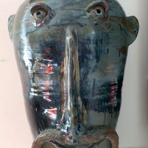 May include: A handcrafted ceramic vase with a face design. The vase features a long nose, eyes, eyebrows, and a mustache. The color is primarily a dark teal with brown and red accents. The vase has a glossy finish and a unique, artistic appearance.