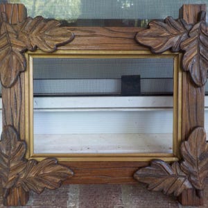 May include: A rectangular wooden picture frame with a gold inner border. The frame is a medium brown color and features carved oak leaves at each corner. The frame is designed to stand upright.