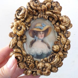 May include: An oval portrait miniature featuring a person wearing a wide-brimmed yellow hat with a blue ribbon, set within a decorative brown frame sculpted with detailed floral and rose motifs that surround the central painted image.