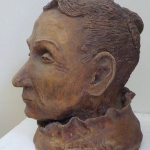 May include: A weathered, brown bust of a person in profile. The sculpture features detailed facial features, including wrinkles and a defined nose. The hair is textured, and the neck is adorned with a ruffled collar. The artwork is likely made of clay or a similar material.