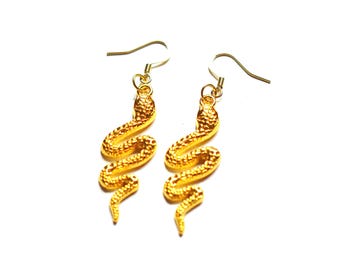 May include: A pair of gold-colored snake earrings. Each earring features a detailed, textured snake design with a curved body and a hook for the ear. The earrings are set against a white background.