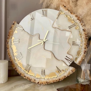 Puede incluir: Un reloj de pared redondo con esfera blanca, números romanos dorados y manecillas doradas. El borde del reloj está adornado con un material texturizado de color ámbar. El reloj tiene un diseño artístico único.