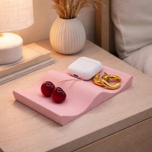 May include: A pink, wavy tray holds a white wireless earbud case, two gold hoop earrings, and two cherries. The tray sits on a wooden nightstand next to a lamp and a vase with dried plants.