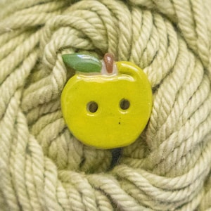 May include: A small, glossy, yellow apple-shaped button with two central holes for attachment. The button features a smooth, rounded surface and a small, green leaf detail at the top, positioned against a background of thick, light green yarn.