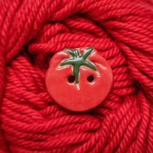 May include: A round, red, tomato-shaped button with a green stem detail and two central holes for attachment. The button features a smooth, glossy finish and is positioned against a background of thick, vibrant red yarn with a visible texture.