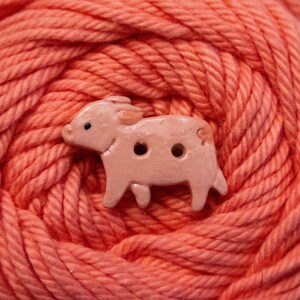 May include: A light peach-coloured button shaped like a pig, featuring a smooth, glossy finish and two small circular holes in the centre for attachment. The pig design includes a small black dot for an eye and a rounded snout, ears, and tail.