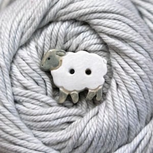 May include: A ceramic button shaped like a sheep with a white body and a grey head and legs. The sheep figure features two holes in the centre for attachment and is positioned against a background of light grey, twisted yarn fibres.