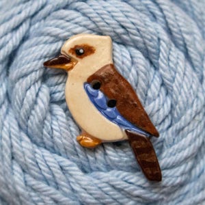 May include: A ceramic button shaped like a kookaburra bird. The bird features a cream-coloured head and chest, a dark brown beak, brown wings with a blue accent stripe, and a brown tail. Two small holes are positioned on the bird's wing area.