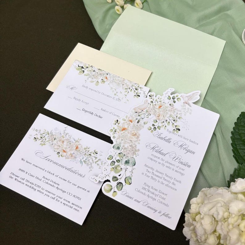 May include: A wedding invitation suite featuring white cardstock with floral watercolor illustrations of white roses and eucalyptus leaves. The set includes a main invitation, an accommodations card, and a response card, all paired with a light green envelope.