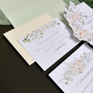 May include: A wedding stationery set featuring white cards with floral illustrations in soft peach and green tones. The response card includes the text Kindly respond by December 12, 2025, Happily Accept, and Regretfully Decline, alongside a pale green envelope.