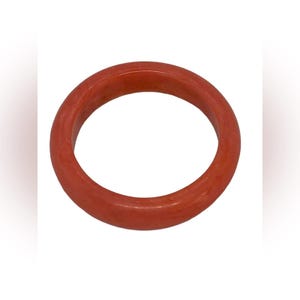 Könnte beinhalten: Ein runder, korallenfarbener Armreif. Das Armband ist ein massives Band in einem rötlich-orangen Farbton. Die glatte Oberfläche reflektiert das Licht und hebt die Farbtiefe hervor. Dieses Accessoire ist ein schlichtes, aber elegantes Schmuckstück.