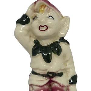 May include: A vintage ceramic figurine of a smiling elf with a white body, red shoes, and a green collar. The elf is wearing a red hat and has black accents. The figurine is in a cheerful pose, with one arm raised.