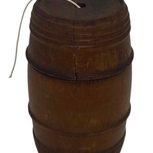 Puede incluir: Un pequeño barril de madera con acabado marrón oscuro. El barril tiene una tapa con un pequeño agujero y una cuerda blanca. Está hecho de duelas de madera y tiene bandas metálicas. Un objeto decorativo.