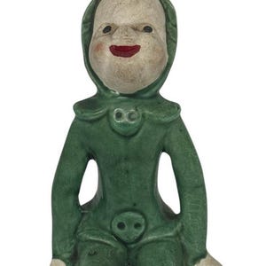 Può includere: Statuetta in ceramica di una figura inginocchiata in tuta verde con cappuccio. La figura ha un viso pallido con occhi neri e una bocca rossa sorridente. La tuta ha due cerchi decorativi sul petto e sull'addome e piedi bianchi.