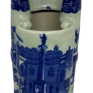 May include: A blue and white ceramic vase with a unique shape. The vase features a detailed blue landscape design on a white background. The design includes buildings, trees, and figures. The vase has a three-part opening at the top.