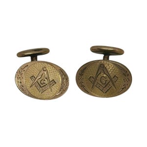 Könnte beinhalten: Ein Paar ovale, goldfarbene Manschettenknöpfe mit strukturierter Oberfläche und einem eingravierten Winkel- und Zirkelsymbol, das in der Mitte mit dem Buchstaben G versehen ist. Die Ränder sind mit einem zarten Blumenmuster verziert.