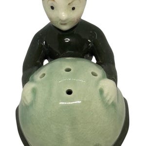 Puede incluir: Figura de cerámica de una persona con un atuendo y gorra verde oscuro, sosteniendo un objeto redondo verde claro con tres agujeros. La figura tiene la piel clara y cabello castaño.