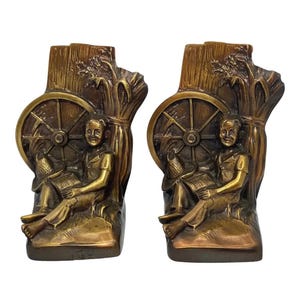 Op de afbeelding: Set van twee boeksteunen in bronskleur. Elke boekensteun toont een zittende figuur bij een wagenwiel en een bundel tarwe. De figuren lezen, en de boekensteunen hebben een rustieke, vintage uitstraling.