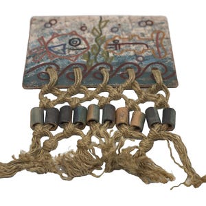 May include: A rectangular wall hanging with a painted aquatic scene. The design features fish and seaweed in blue, red, and green. It is attached to a macrame design with wooden beads and tassels, creating a nautical theme.