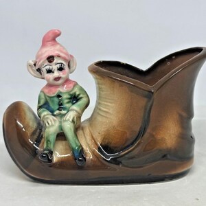 May include: A brown ceramic shoe-shaped planter featuring a small elf figure with a pink hat and green clothing sitting on the front. The glazed finish transitions from dark brown at the base to a lighter tan tone on the upper part of the shoe.
