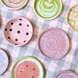 Può includere: Una collezione di piccoli piattini rotondi in ceramica con vari disegni, tra cui motivi di latte art, pois neri su fondo rosa, una spirale rosa, un motivo a fetta di anguria e un piattino rosa con pois bianchi su un tessuto a quadri.