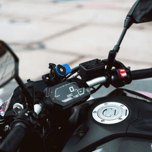Puede incluir: Primer plano del salpicadero digital de una motocicleta, que muestra la velocidad y otros datos. El salpicadero negro tiene detalles en blanco y rojo. Un soporte azul para teléfono está fijado al manillar. El depósito de combustible es visible.