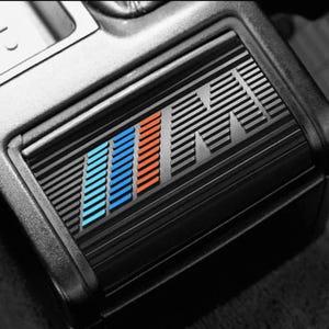 Pode incluir: Detalhe interior de carro preto com um emblema "M" estilizado. O emblema apresenta listras azuis e laranja sobre um fundo de linhas horizontais. O design é moderno e elegante, com uma aparência texturizada.